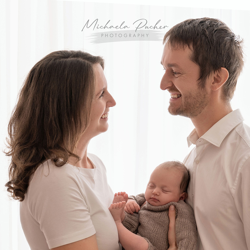 Ein Paar steht einander lächelnd gegenüber, während der Vater ein schlafendes Baby im gestrickten Outfit im Arm hält; weiches Gegenlicht fällt durch einen weißen Vorhang.