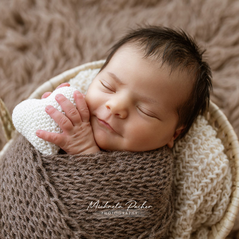Schlafendes Neugeborenes liegt eingewickelt in braune Strickstoffe in einem Korb und hält ein kleines weißes Häkelherz in der Hand.