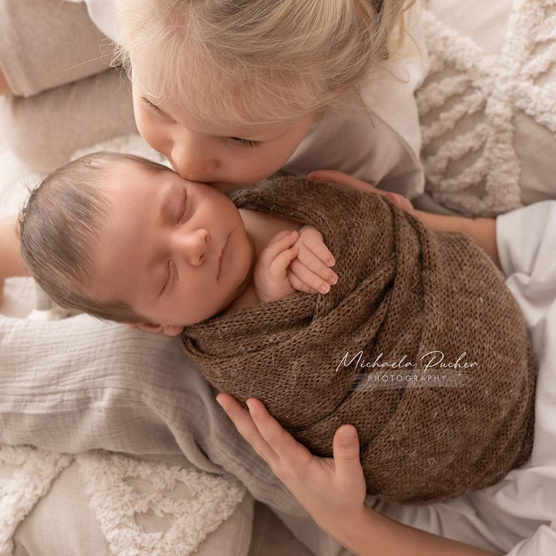 Ein kleines Mädchen küsst vorsichtig das schlafende, eingewickelte Neugeborene auf die Stirn; beide liegen auf weichen, neutralen Stoffen.