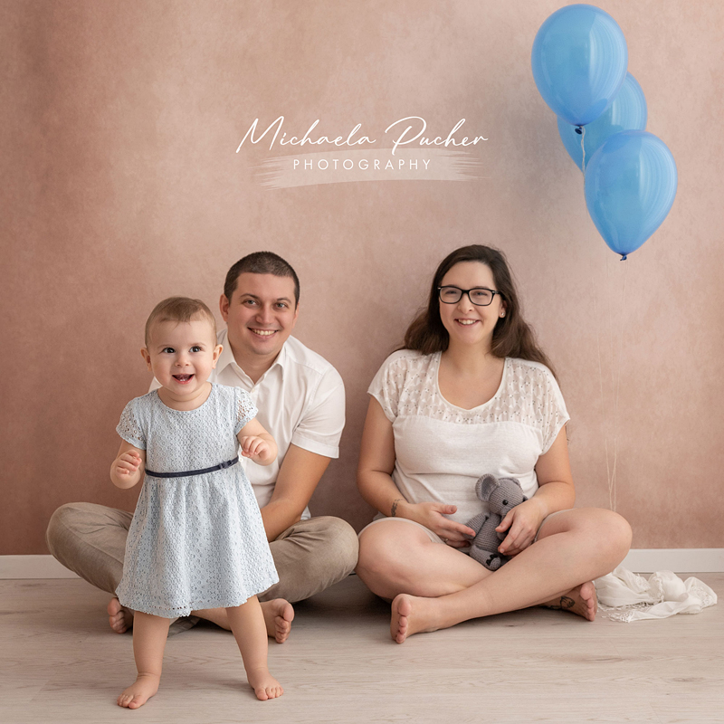 Eine Familie sitzt entspannt auf dem Boden vor roséfarbenem Hintergrund; das kleine Mädchen läuft fröhlich im Vordergrund, während Mama und Papa dahinter sitzen und lächeln. Rechts schweben drei blaue Luftballons.