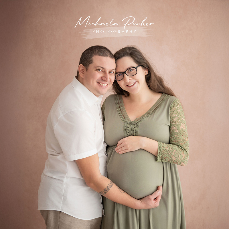 Ein Paar steht eng beisammen vor einem roséfarbenen Hintergrund; er hält liebevoll den Babybauch seiner schwangeren Partnerin, beide lächeln in die Kamera.