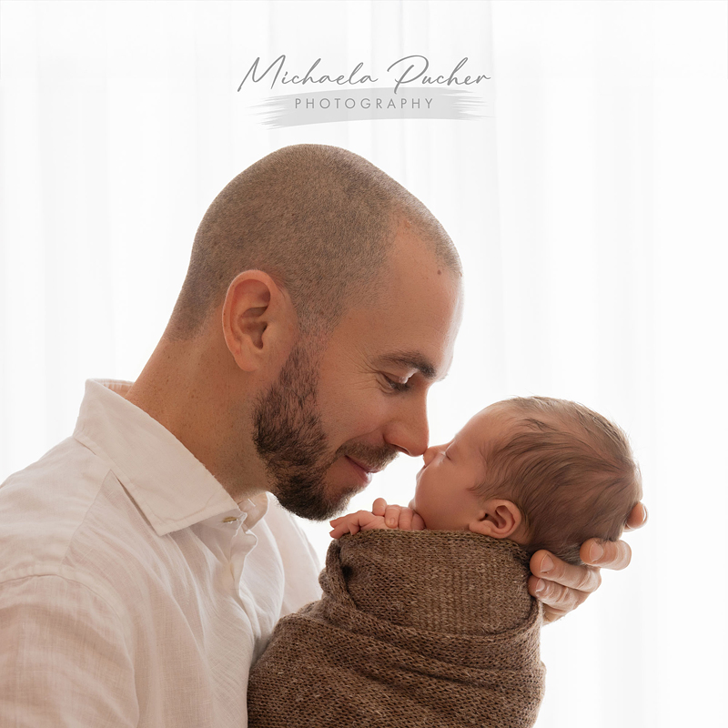 Ein Vater hält sein schlafendes, eingewickeltes Neugeborenes nah am Gesicht und berührt mit der Nase sanft dessen Stirn; weiches Gegenlicht fällt durch einen weißen Vorhang.