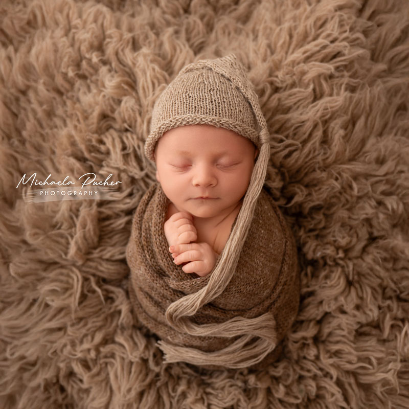 Ein schlafendes Baby liegt eingewickelt in braune Strickstoffe auf einem flauschigen Fell; es trägt eine gestrickte Mütze mit langen Bändern und hält die Hände leicht vor der Brust.