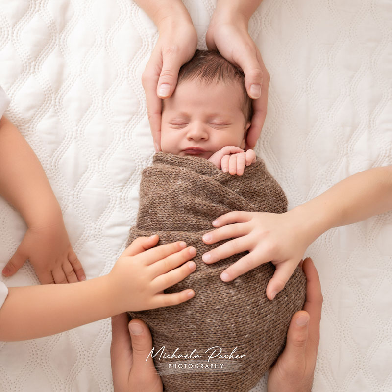 Ein schlafendes, eingewickeltes Neugeborenes liegt auf einer weißen, strukturierten Decke; mehrere Kinderhände berühren sanft den Körper, während zwei erwachsene Hände schützend den Kopf halten.