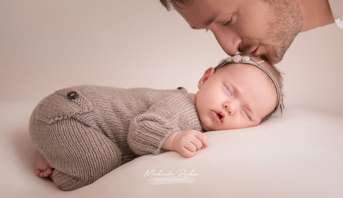 Schlafendes Neugeborenes in einem gestrickten beige-grauen Overall, während der Vater seinem Baby sanft einen Kuss auf die Stirn gibt.