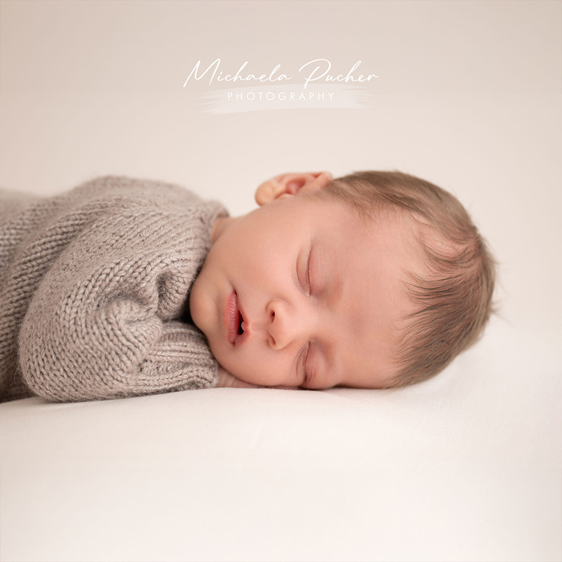 Ein schlafendes Baby liegt seitlich auf einem weichen Untergrund; es trägt einen gestrickten beigefarbenen Overall und hat den Kopf entspannt auf die Händchen gelegt.