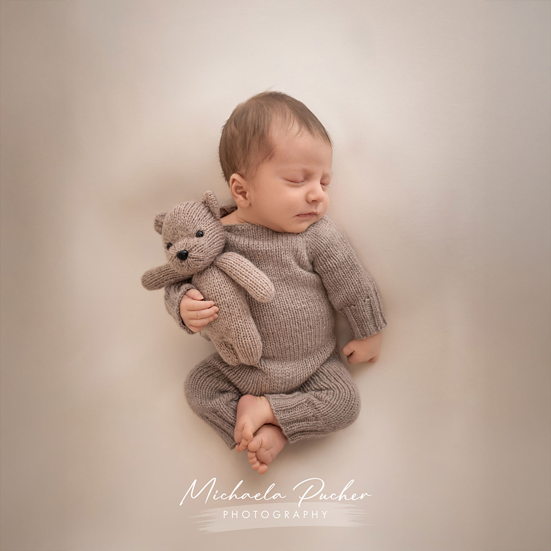 Ein schlafendes Baby liegt in einem gestrickten braunen Outfit auf dem Rücken und hält ein passendes gestricktes Bärchen im Arm.