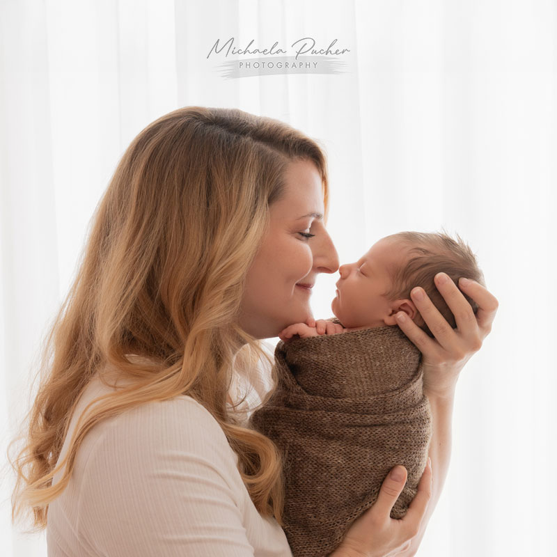 Eine Mutter hält ihr schlafendes, eingewickeltes Neugeborenes nah am Gesicht, beide berühren sich sanft mit der Nase; weiches Gegenlicht fällt durch einen weißen Vorhang.