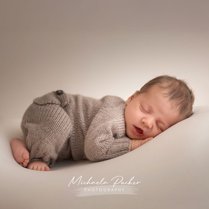 Ein schlafendes Baby liegt seitlich in einem gestrickten beigefarbenen Overall, den Kopf entspannt auf die Hände gelegt; die Lippen sind leicht geöffnet.