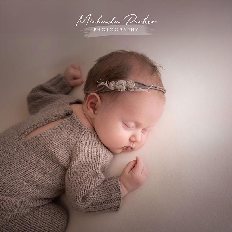 Ein schlafendes Baby liegt auf der Seite in einem gestrickten, beigefarbenen Outfit; ein zartes Stirnband mit kleinen Stoffblüten schmückt den Kopf.