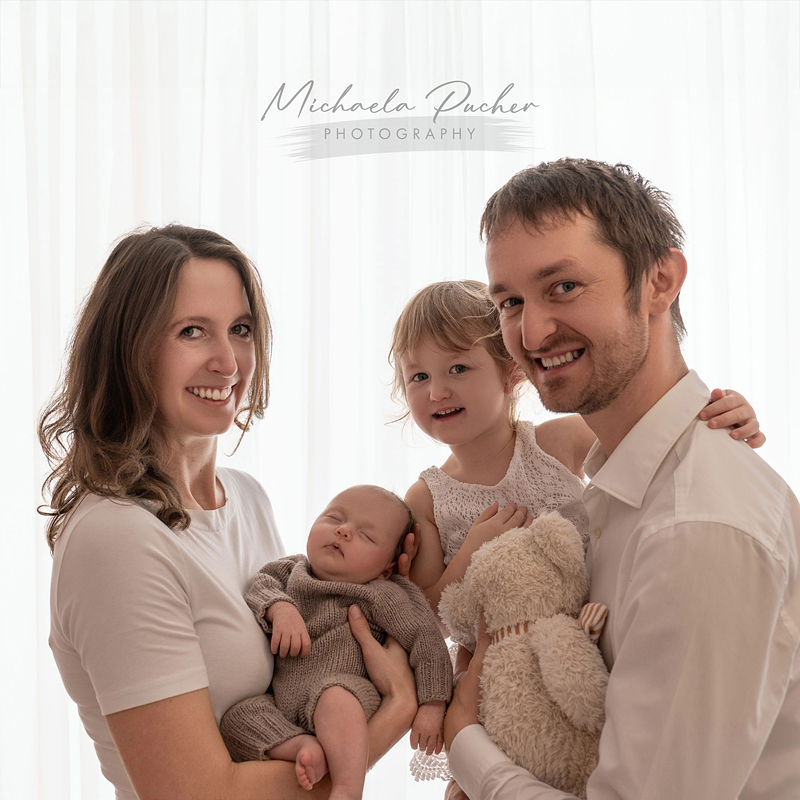 Eine vierköpfige Familie steht eng beisammen vor einem hellen Vorhang; die Mutter hält das schlafende Baby, während der Vater die große Schwester im Arm hat, die einen Teddybären hält.