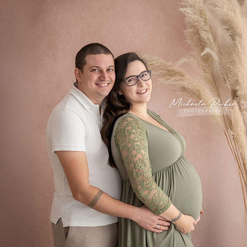 Ein Paar steht eng beisammen vor einem warmen roséfarbenen Hintergrund; der Mann umarmt die schwangere Frau von hinten und hält liebevoll ihren Babybauch, beide lächeln in die Kamera.