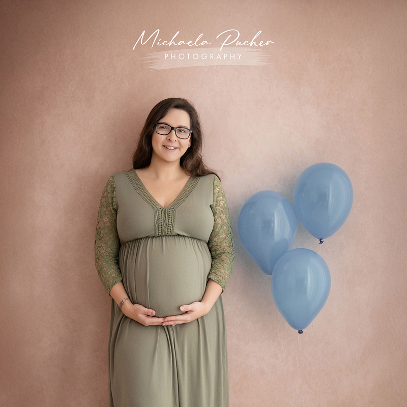 Eine schwangere Frau steht vor einem roséfarbenen Hintergrund, trägt ein grünes Kleid mit Spitzenärmeln und hält ihren Babybauch; daneben schweben drei blaue Luftballons.