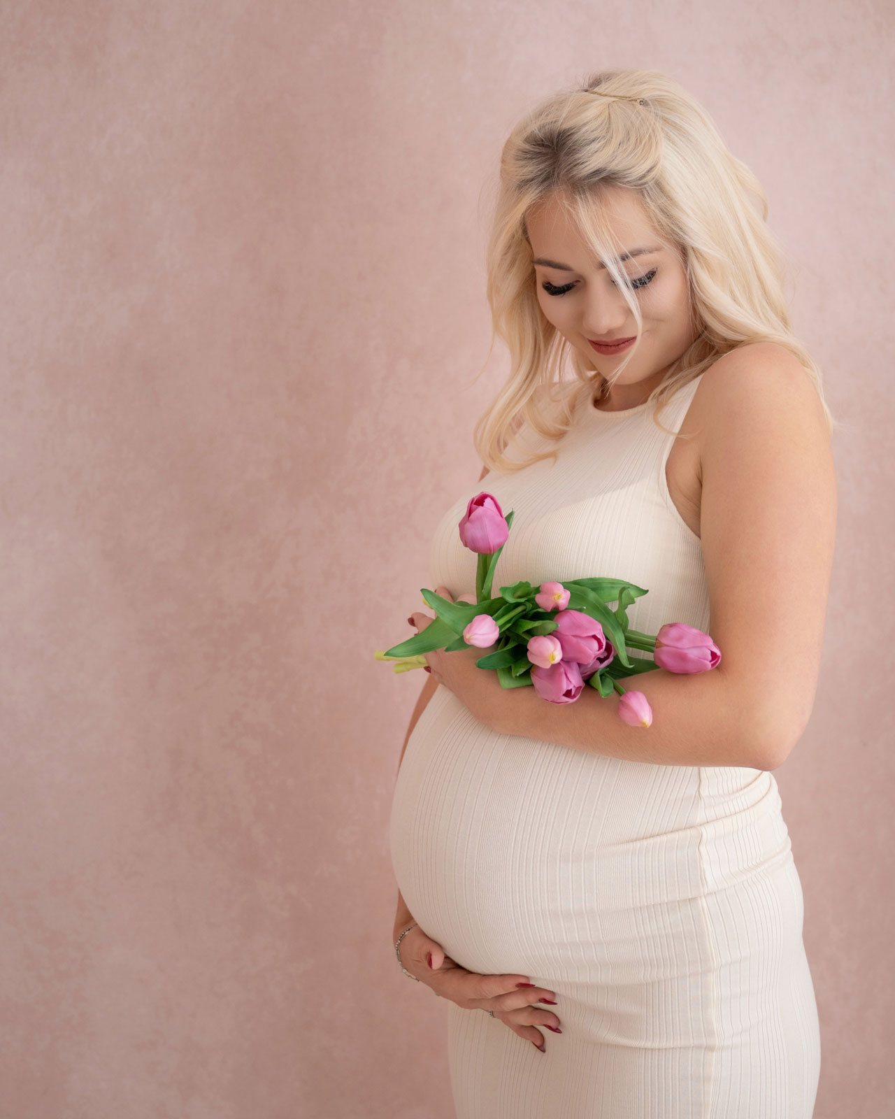 Schwangere Frau im cremefarbenen Kleid hält einen Tulpenstrauß und eine Hand am Babybauch