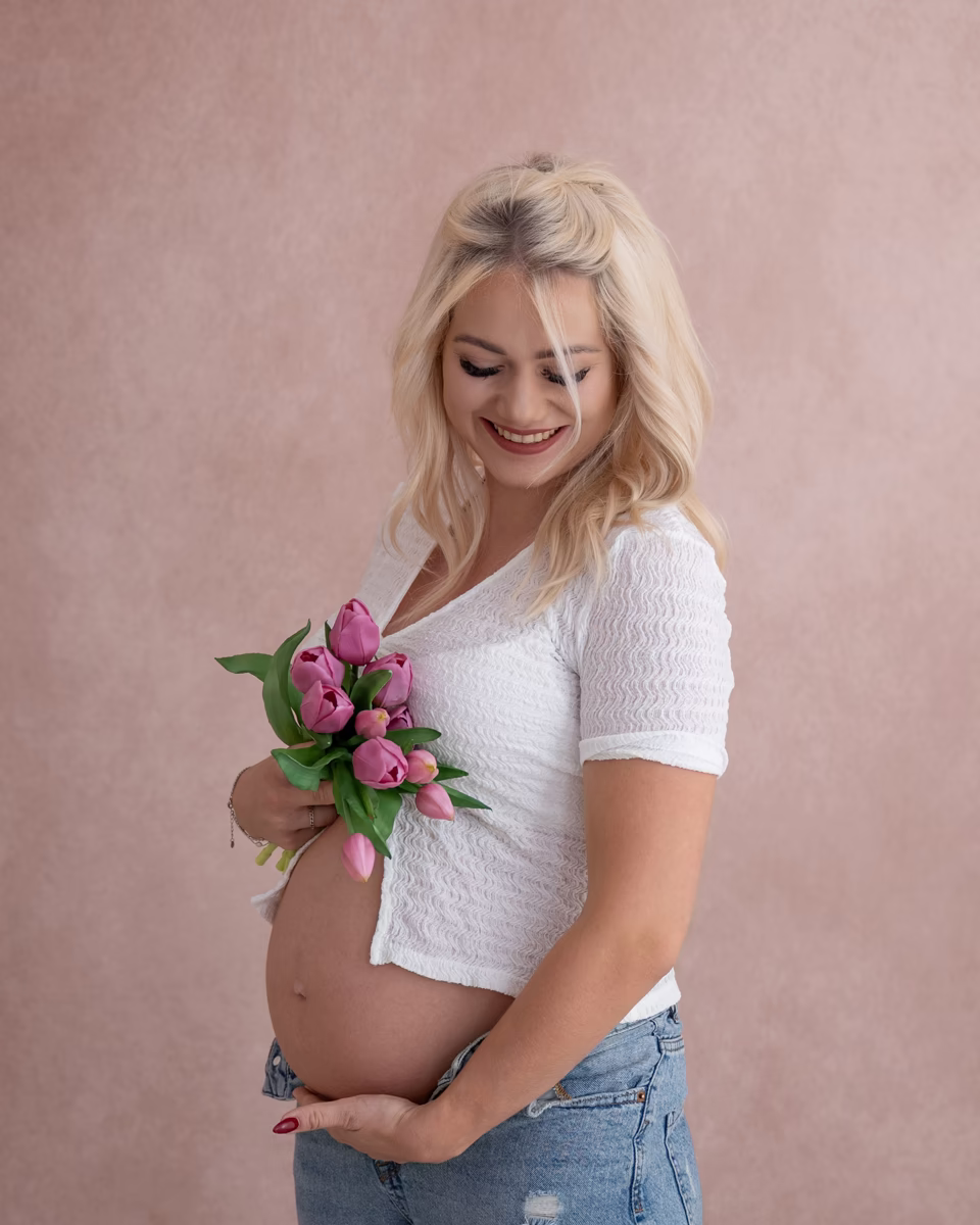 Schwangere Frau in weißem Shirt und Jeans hält einen Strauß rosa Tulpen an ihren Babybauch