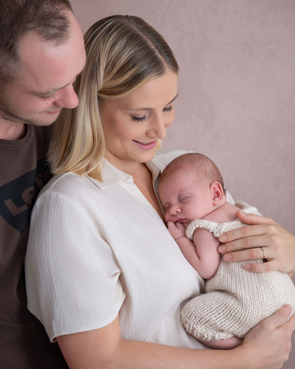 Eltern halten ihr schlafendes Neugeborenes im Arm, der Papa schaut über die Schulter der Mama